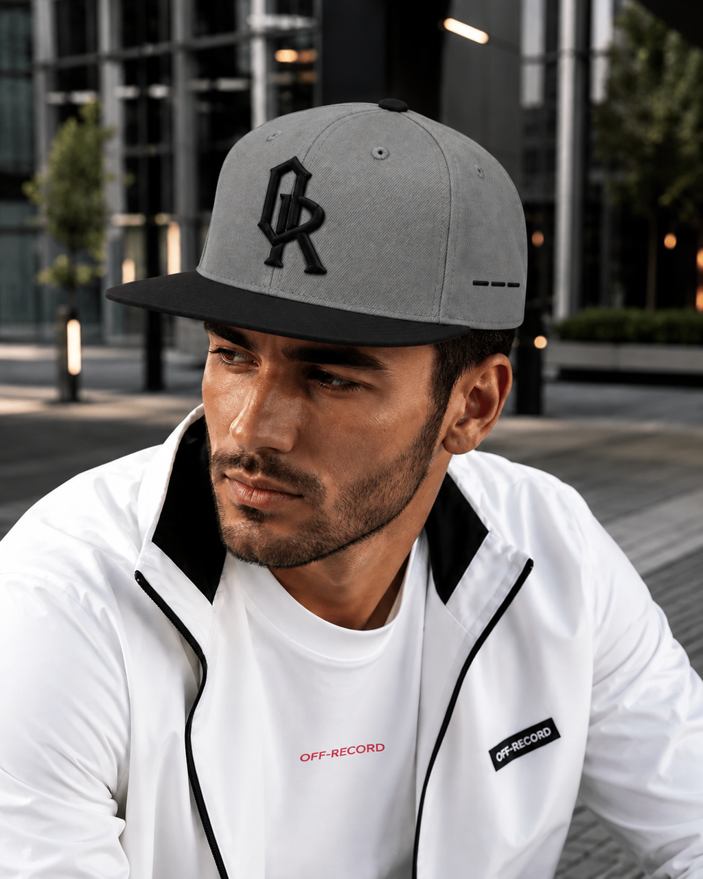 A man wearing a grey and black Off-Record snapback cap and a white zip-up jacket with black trim in an outdoor urban setting.