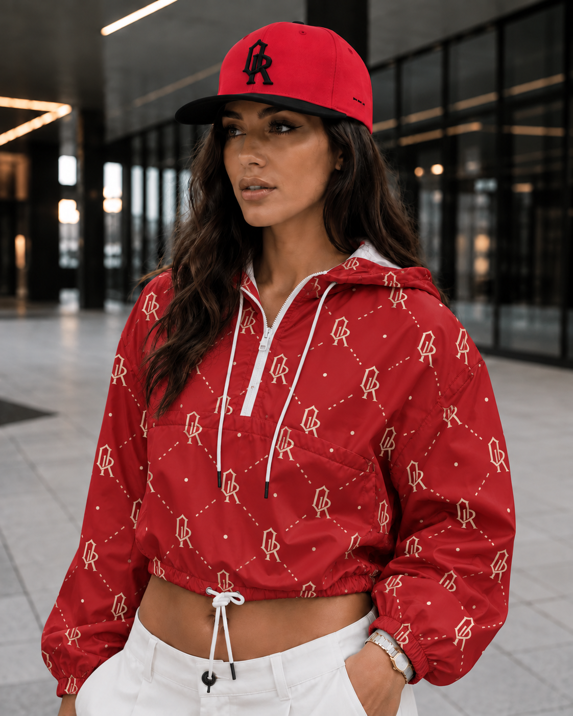 A woman wearing a red cropped hooded jacket with a white and red geometric monogram pattern, a matching red baseball cap with a black logo, and white drawstring pants, standing in a modern indoor setting.