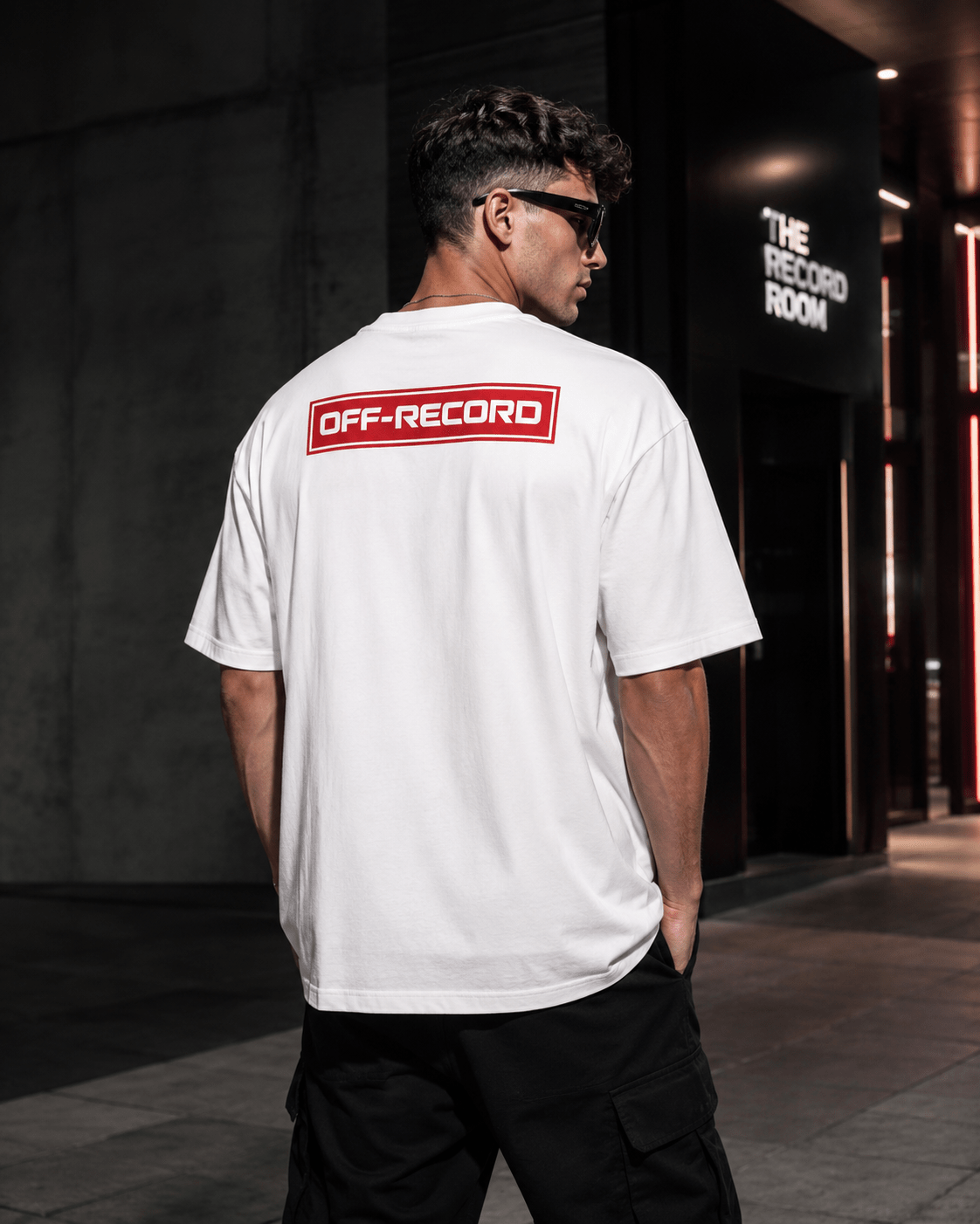 Person wearing a white Off-Record reverse stamp tee logo t-shirt with 'OFF-RECORD' logo on the back, standing in an urban setting.